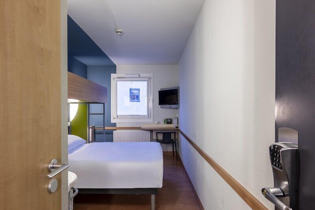 A compact hotel room with a bed, a desk, a chair, a television, and a window.