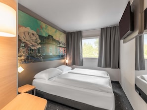 Habitación de hotel con cama, escritorio, ventana con cortinas, televisor montado en la pared y un cuadro.