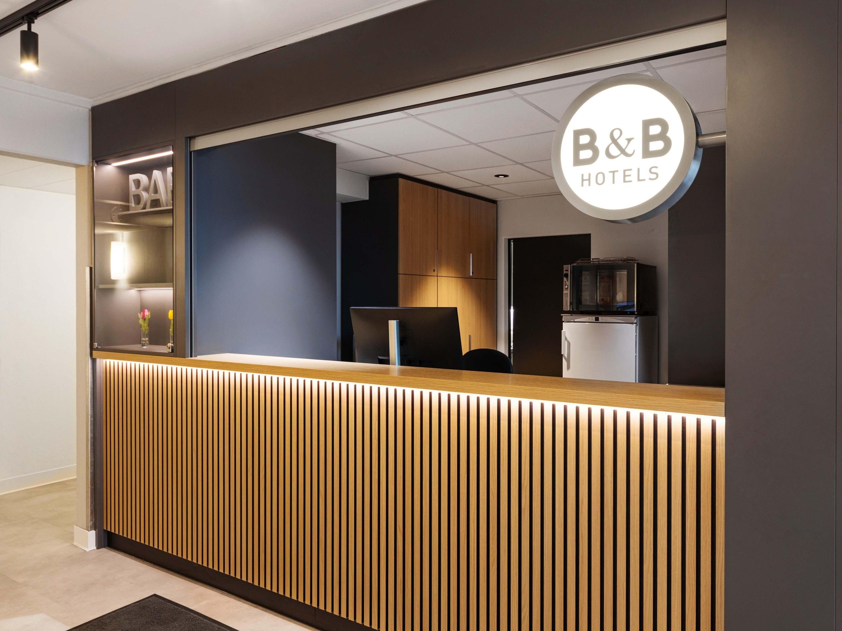 Un comptoir d’accueil au design moderne, une enseigne indiquant qu’il fait partie du groupe B&B Hotels, et un aperçu de l’intérieur, notamment du comptoir et d’un réfrigérateur.