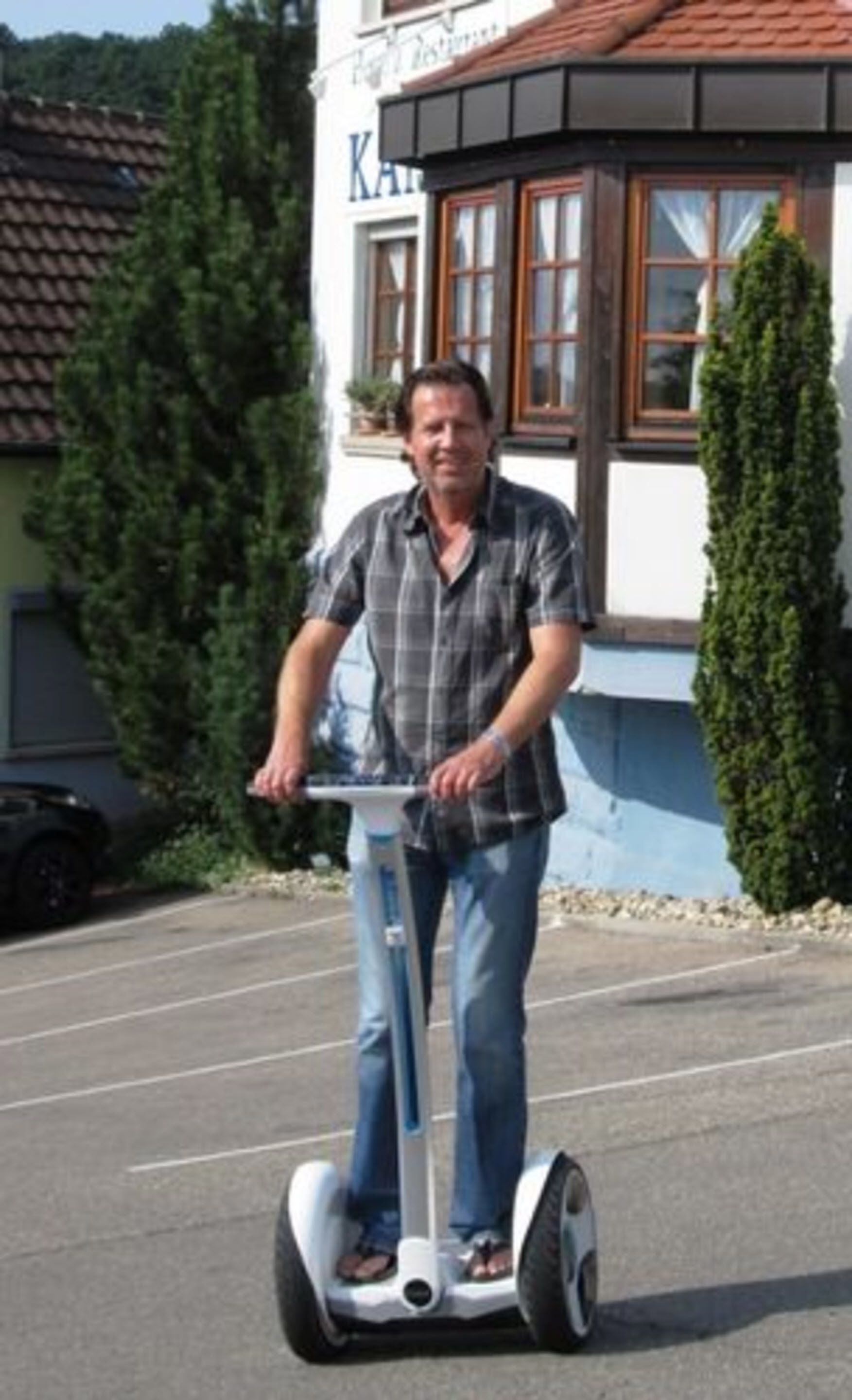 Eine Person fährt auf einem Segway über eine gepflasterte Fläche vor einem Gebäude mit einem Ziegeldach und einem Fenster mit Vorhängen.