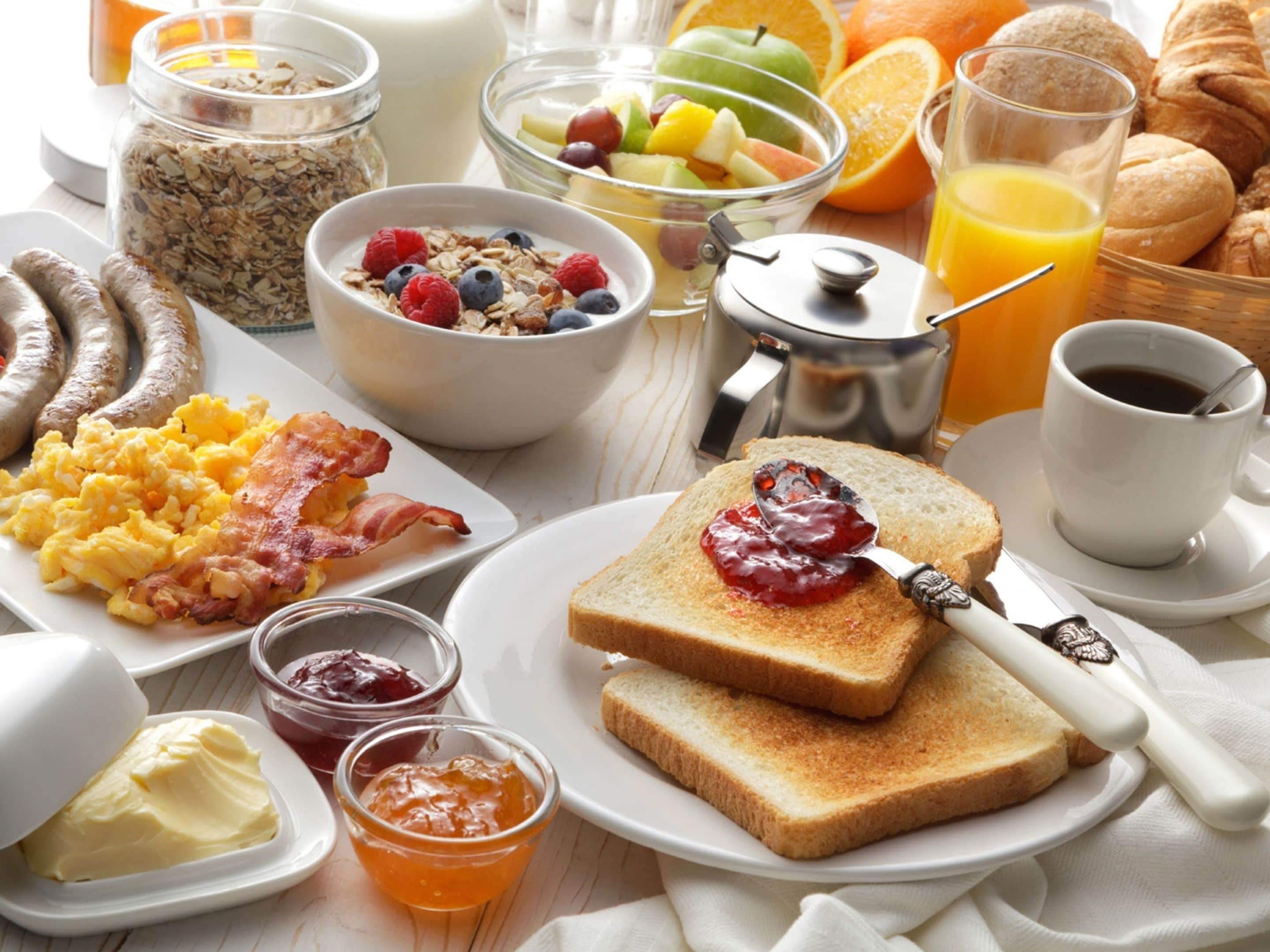 Una mesa de desayuno con diversos alimentos, incluyendo tostadas, huevos revueltos, salchichas y un bol de fruta.