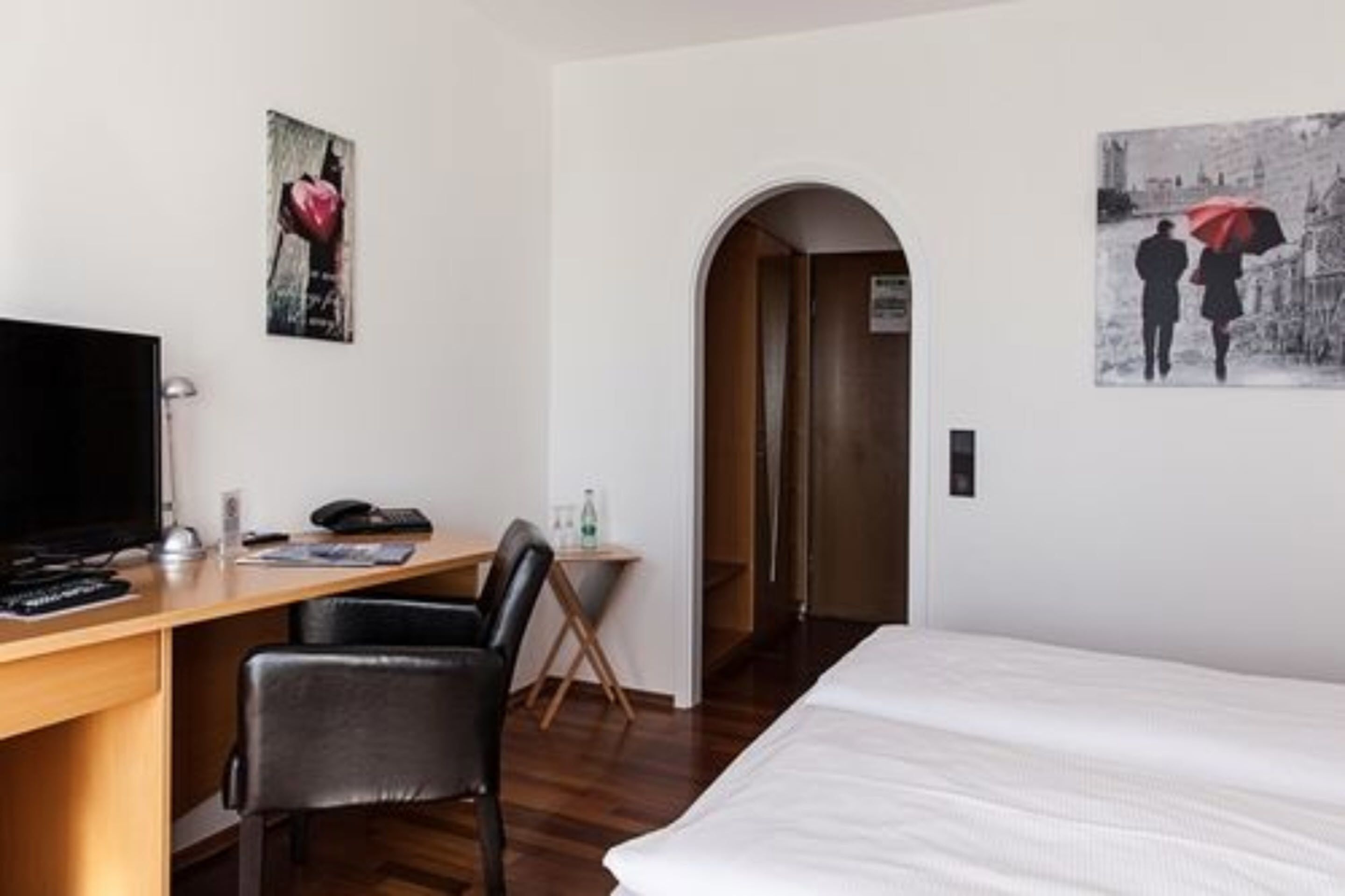 A hotel room with a bed, a desk with a computer, a chair, and two paintings on the wall.