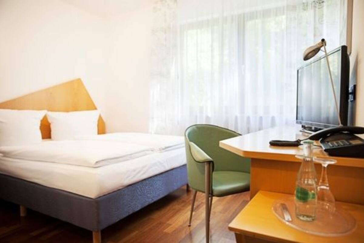 A hotel room with a bed, a desk with a lamp, a chair, and a bottle on the desk.