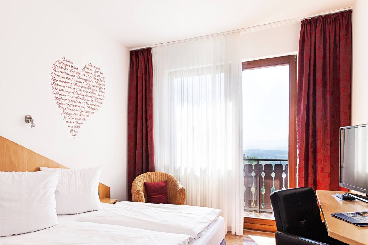 A hotel room with a bed, a desk with a computer, a chair, and a balcony with a view.