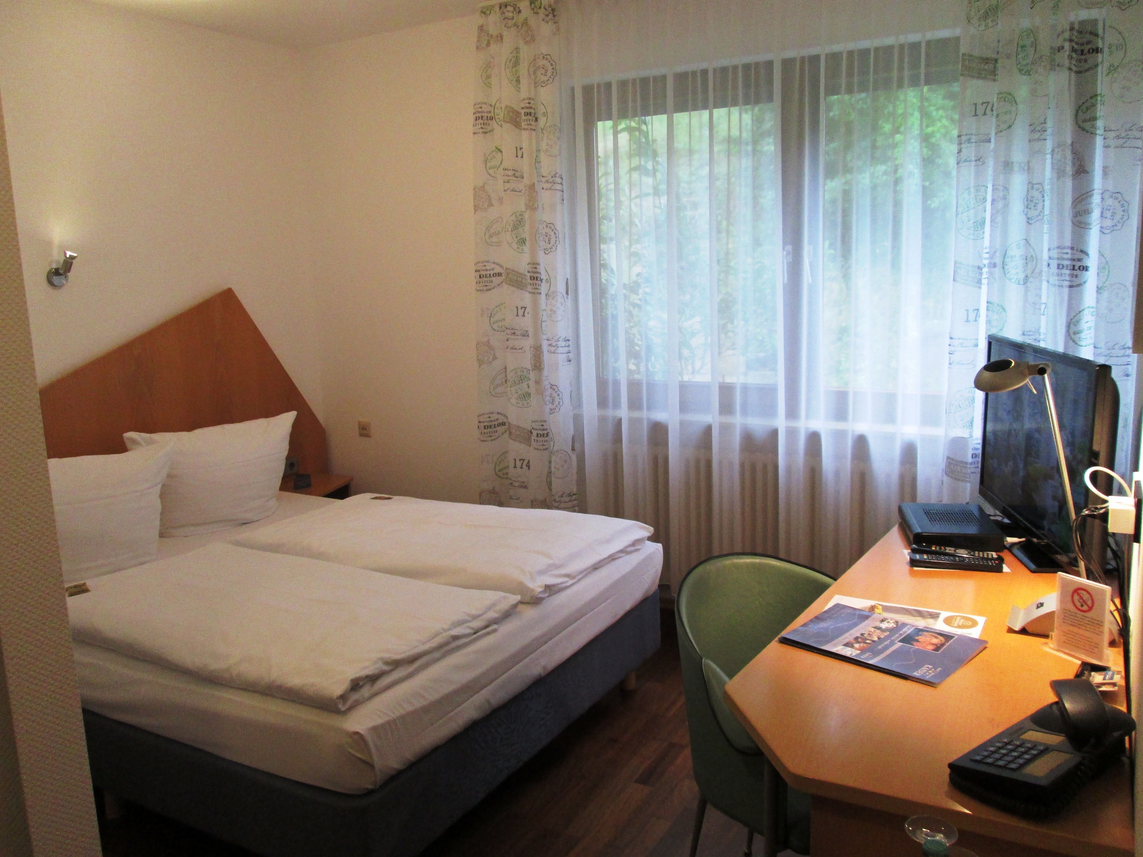 Quarto de hotel com duas camas, uma escrivaninha com computador e telefone, e uma janela com cortinas leves.