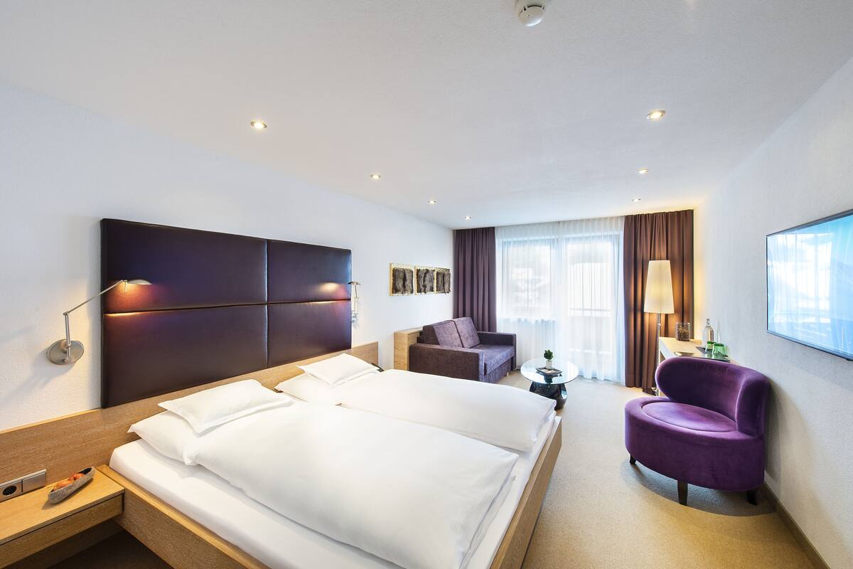 A hotel room with a large bed, a sofa, a purple armchair, a small table, and a window with curtains.