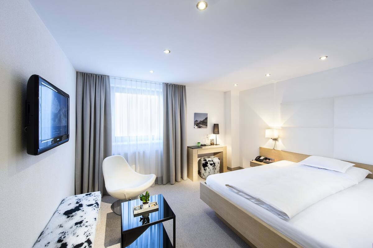 A hotel room with a bed, a desk with a flat-screen TV, a chair, a bedside table with a lamp, and a window with curtains.