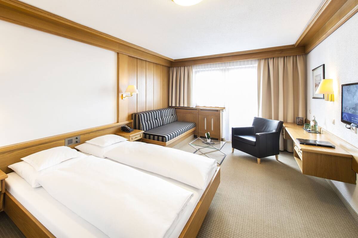 A hotel room with a large bed, a sofa, a desk with a chair, a TV, and a window with curtains.