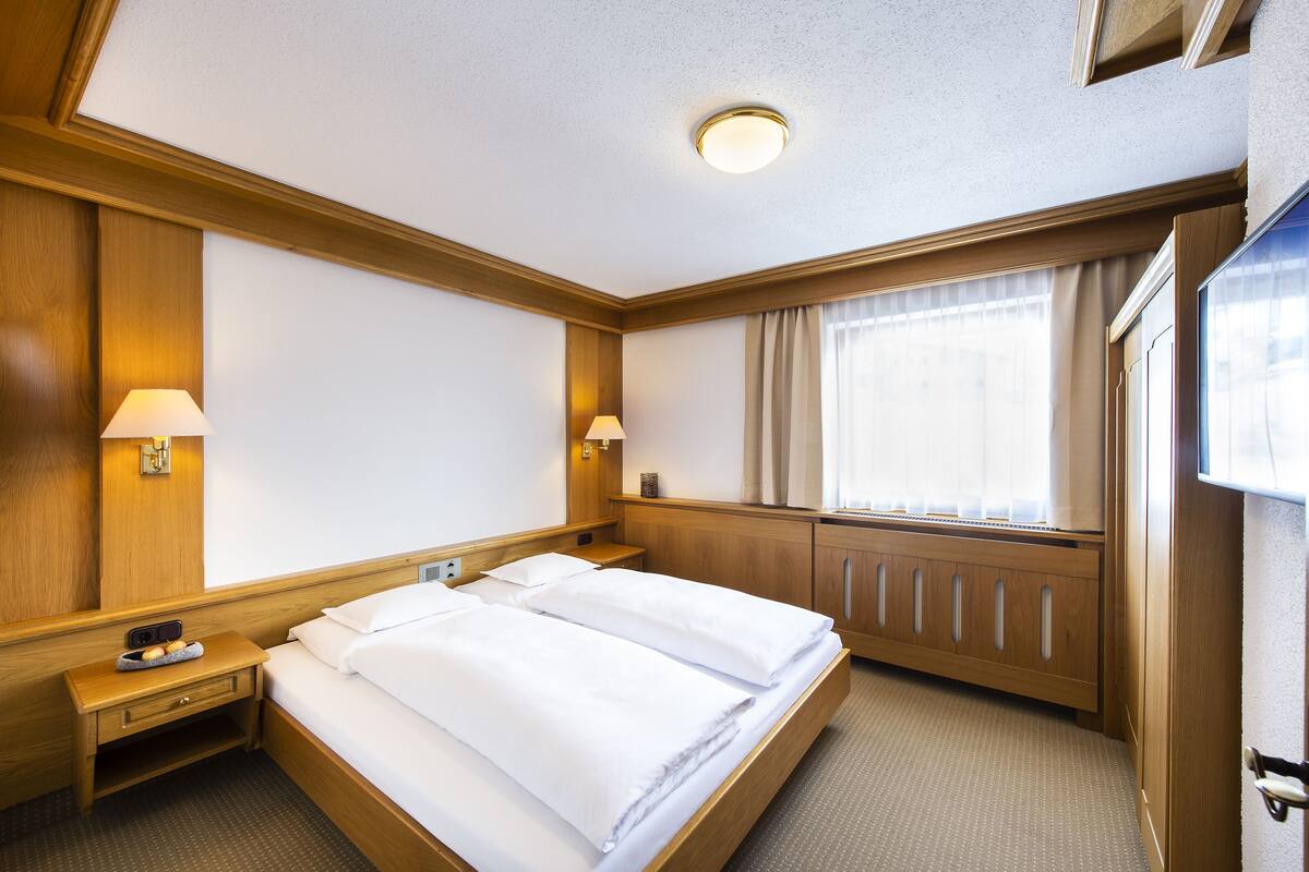 A hotel room with a double bed, wooden headboard, bedside lamps, a nightstand, a window with curtains, and a radiator.