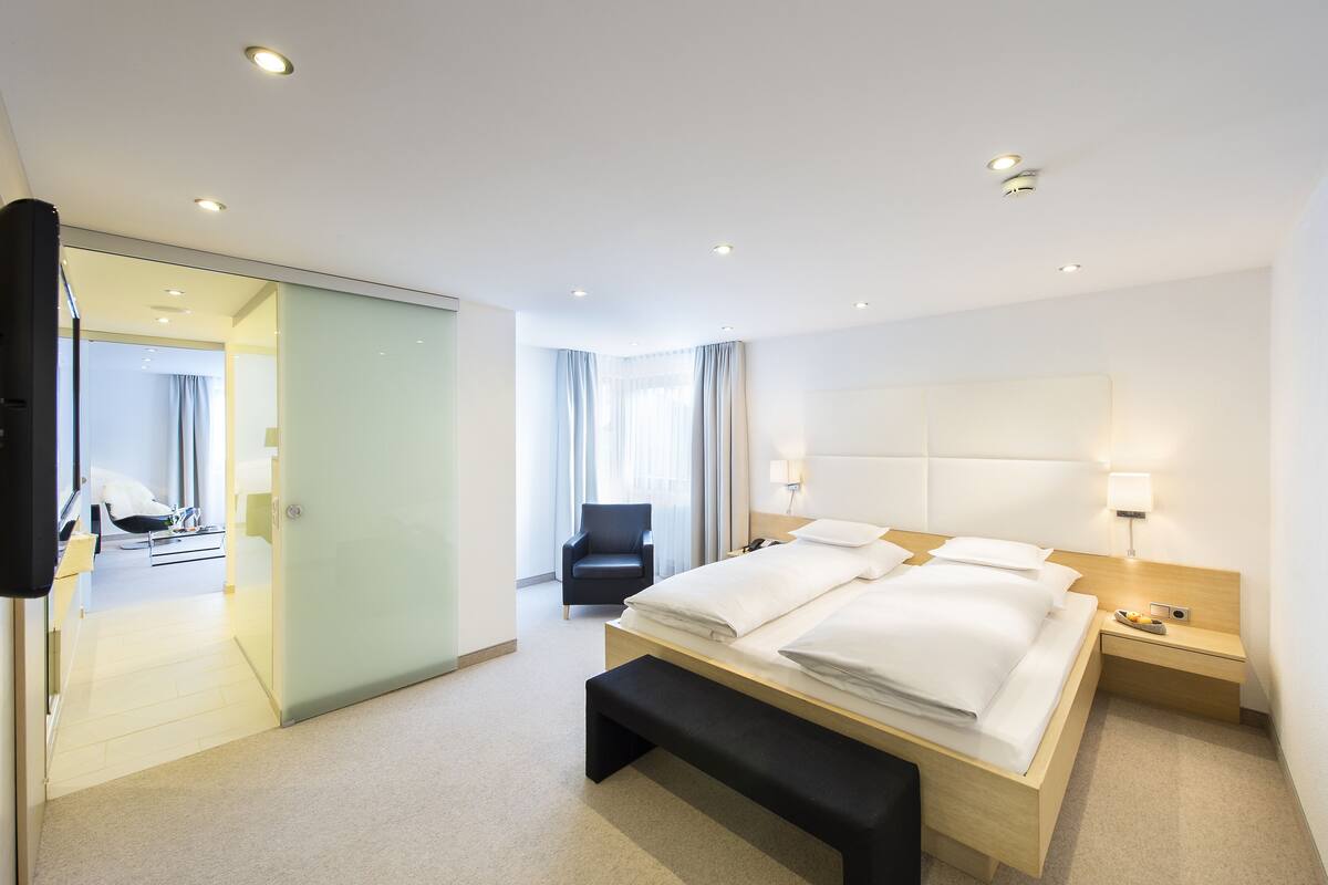A modern hotel room with a large bed, bedside tables, a bench, a TV, and a glass door leading to a bathroom.