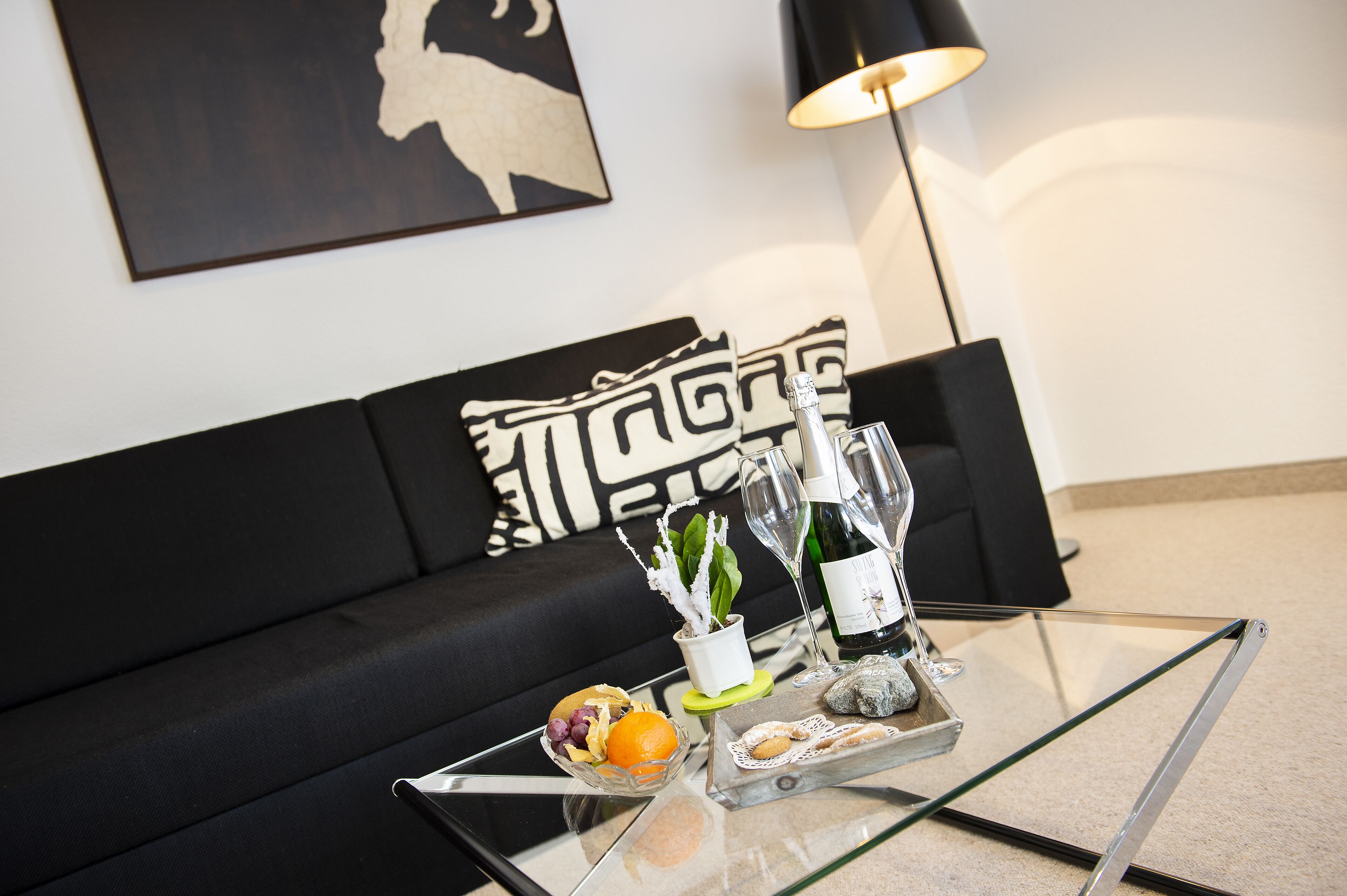 A glass coffee table with a tray of snacks, a bottle, and a glass of water in a modern living room with a black sofa and a lamp.