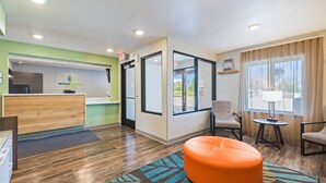 Zona de recepción con un escritorio de madera, mostrador de recepción y un cartel de WoodSpring Suites. Hay dos sillas y una mesita con una lámpara. El piso es de madera y hay una alfombra colorida.