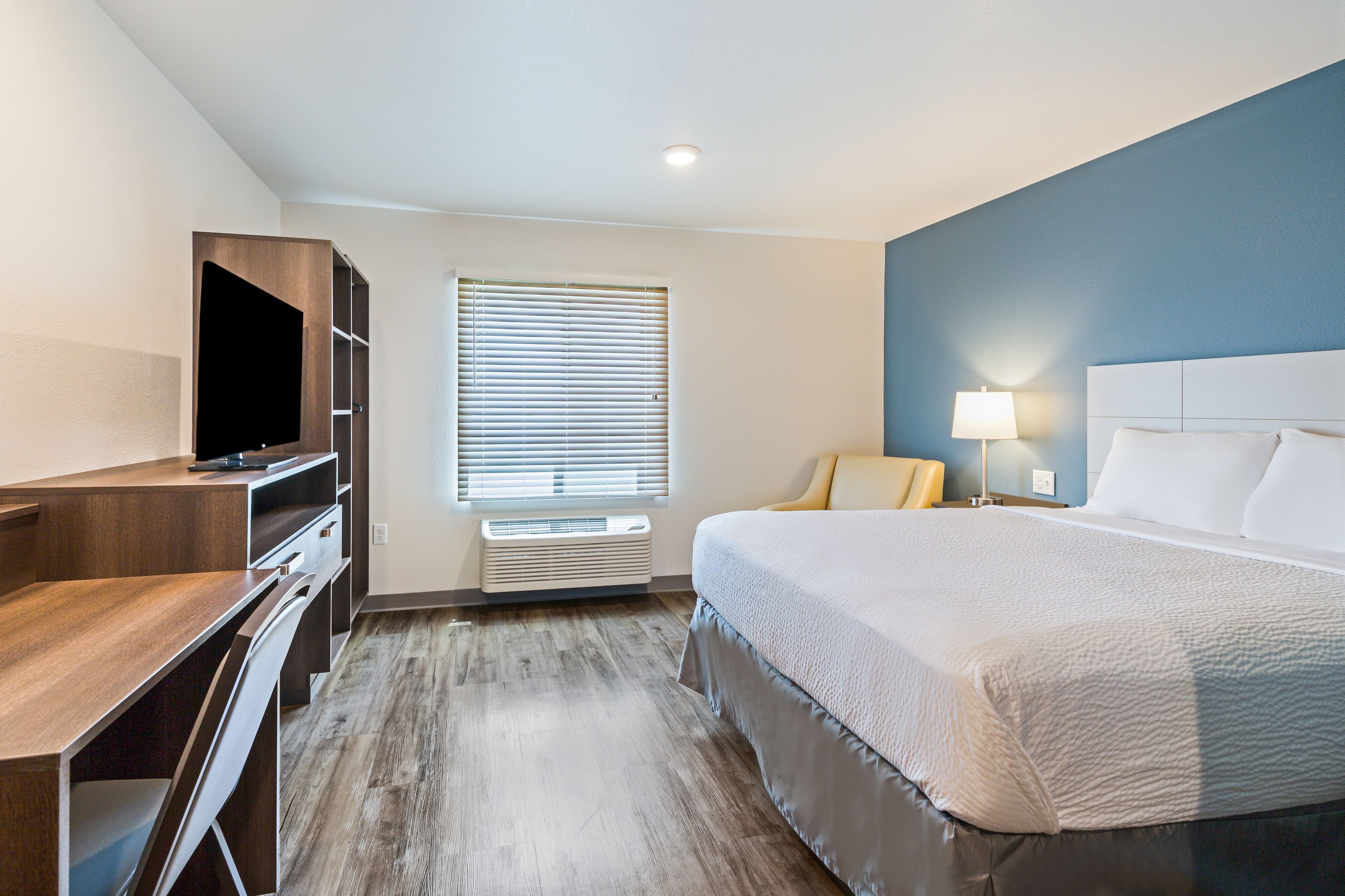 A hotel room with a bed, a desk, a TV, a window with blinds, and a lamp.