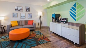 Una habitación de hotel moderna con un sofá gris, un otoman naranja y una alfombra estampada. Hay cuadros enmarcados en la pared, una lámpara y un minibár con cafetera.
