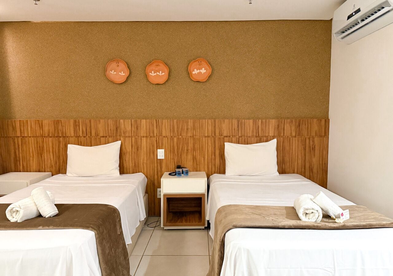 A hotel room with two beds, a wooden headboard, a nightstand with a lamp, and a wall-mounted air conditioner.