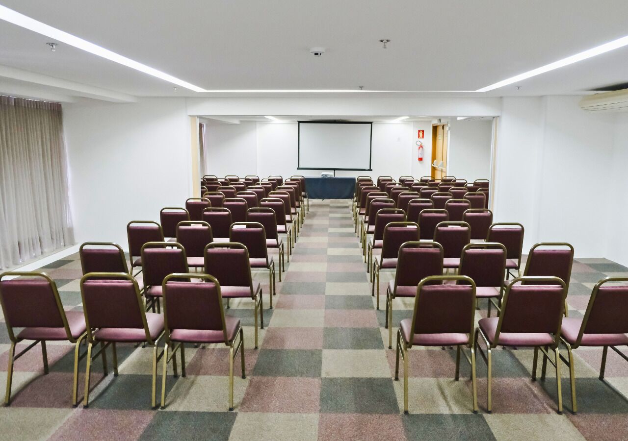 Une salle de conférence avec des rangées de chaises, un écran de projection et un tapis à carreaux.