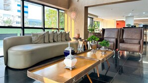 Een moderne hotelentree met een bank, twee houten tafels en een bruine lederen fauteuil.