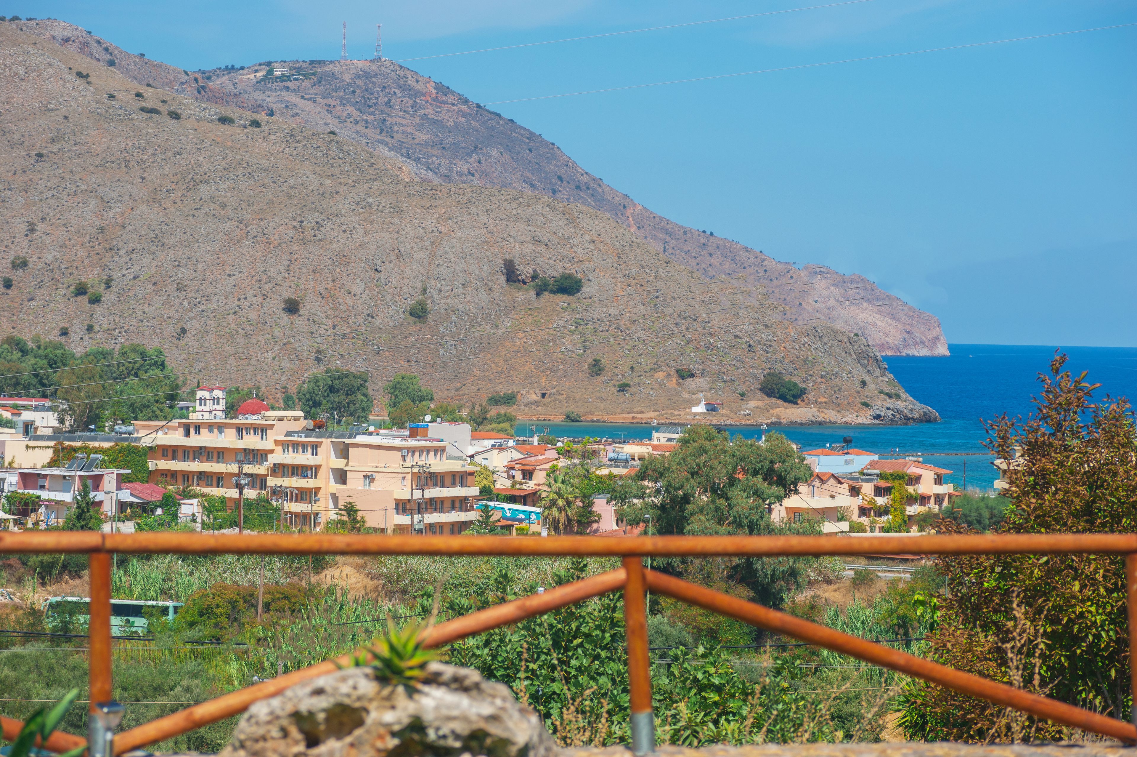 A coastal town with buildings on a hillside, a clear view of the sea, and a metal railing in the foreground.