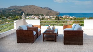 Eine Terrasse mit Korbmöbeln und Blick auf eine Küstenstadt und Berge.