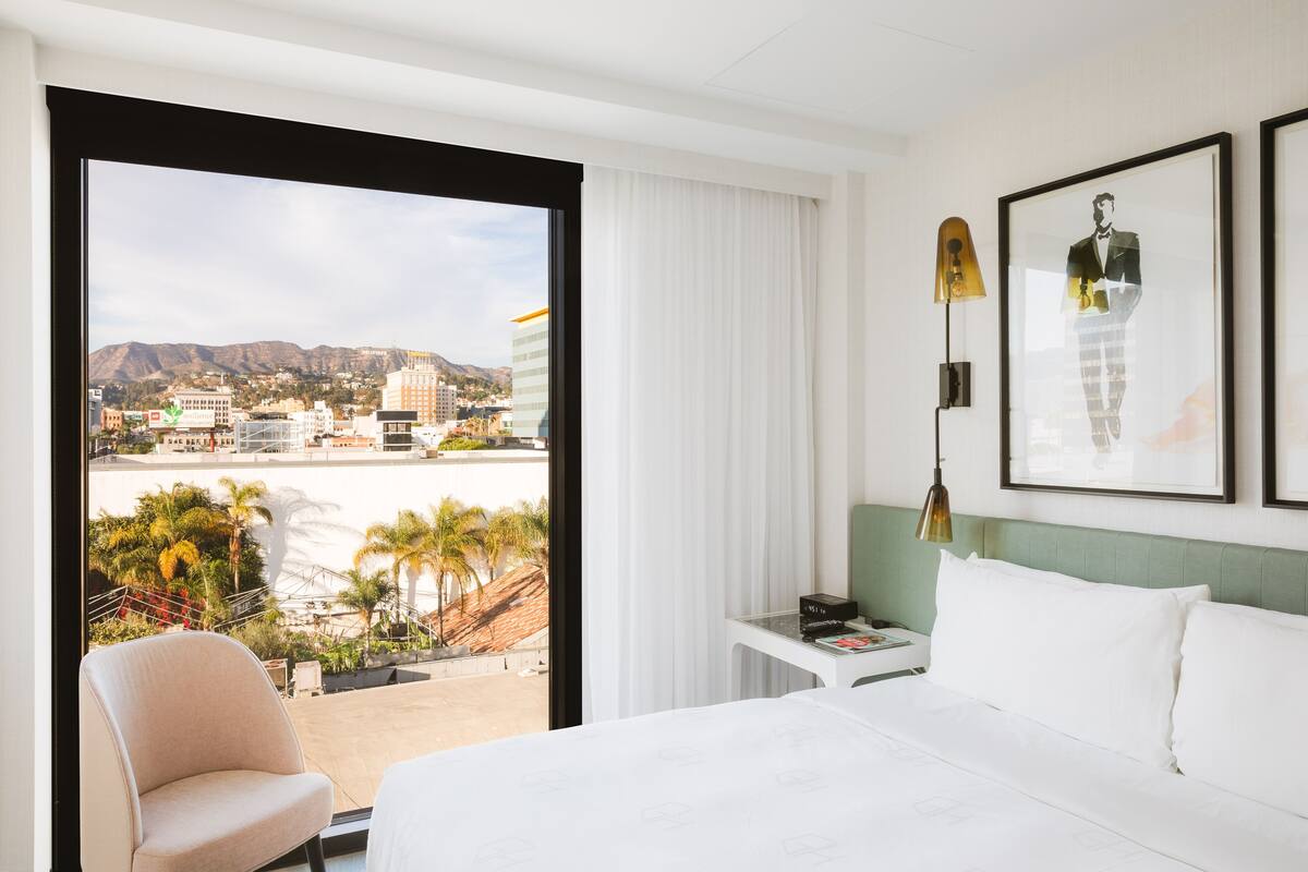 Ein Hotelzimmer mit einem großen Bett, einem Schreibtisch, einem Sessel, einem Nachttisch und Blick auf eine Stadtlandschaft und Berge.