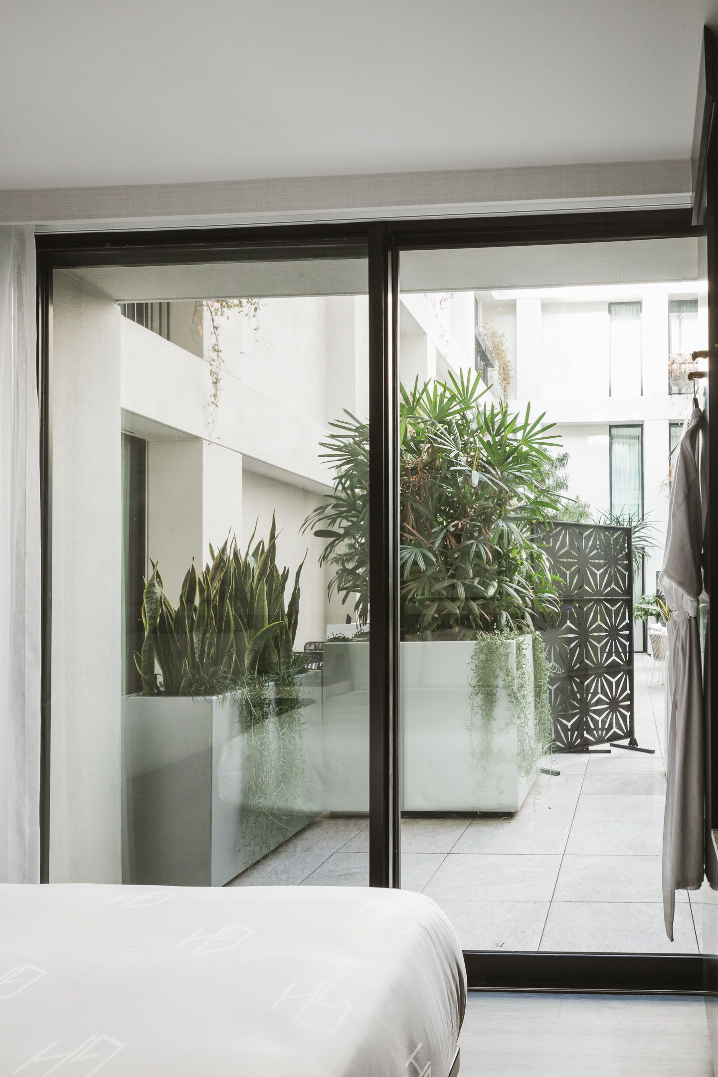 A modern bedroom with a large sliding glass door leading to a balcony with potted plants and a geometric fence.