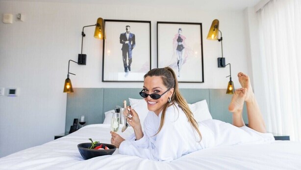 A person lying on a bed with a bowl of fruit and two glasses of champagne, in a room with a modern design, two framed pictures on the wall, and two wall-mounted lamps. - The Godfrey Hotel Hollywood (Los Angeles)