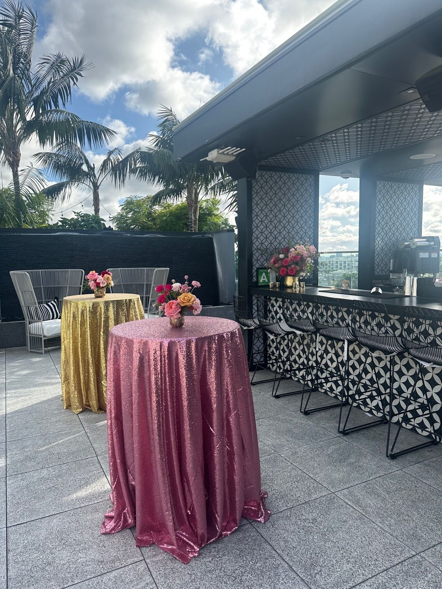 An outdoor seating area with tables covered in gold and pink fabric, chairs, and a bar area with patterned stools.