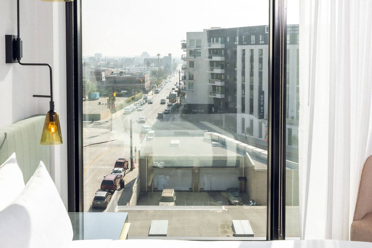 Ein Hotelzimmer mit großem Fenster und Blick auf die Stadt, einem Bett mit weißer Bettwäsche und einer modernen Lampe.