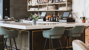 Una zona de bar con mostrador de mármol, varias estanterías llenas de botellas, un ordenador portátil y tres taburetes.