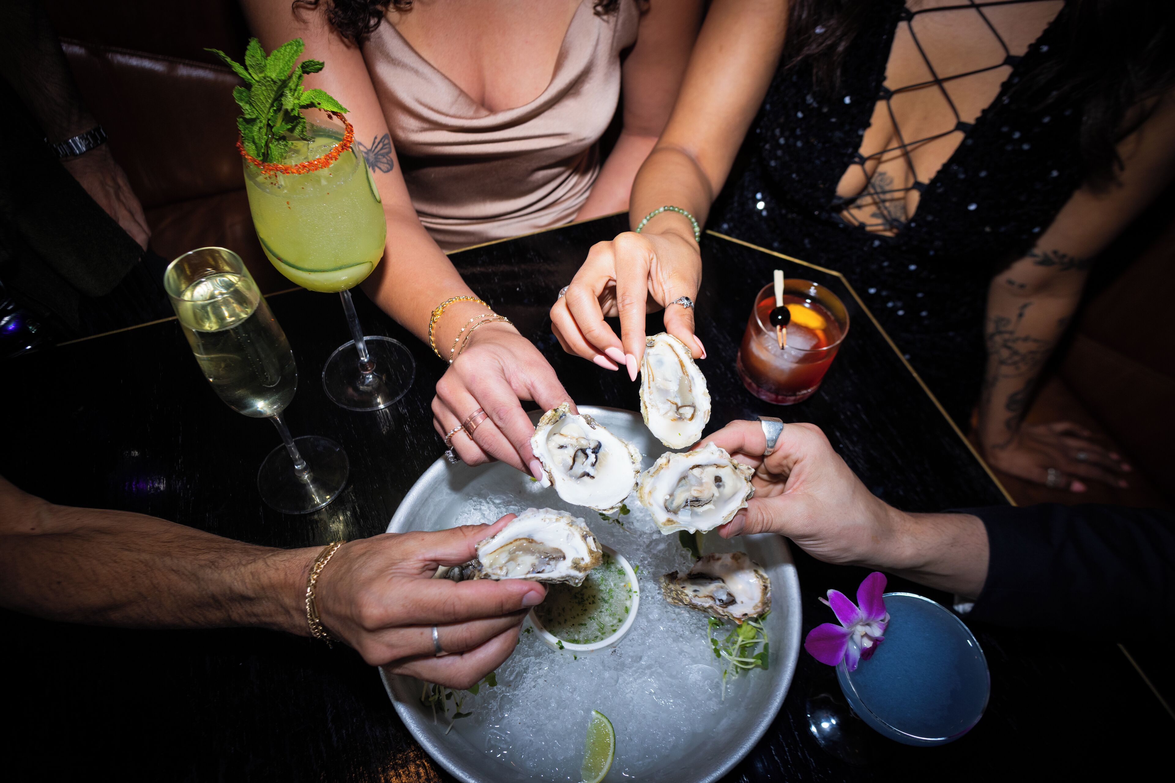 A group of people at a restaurant table with oysters, drinks, and a small bowl of greens.