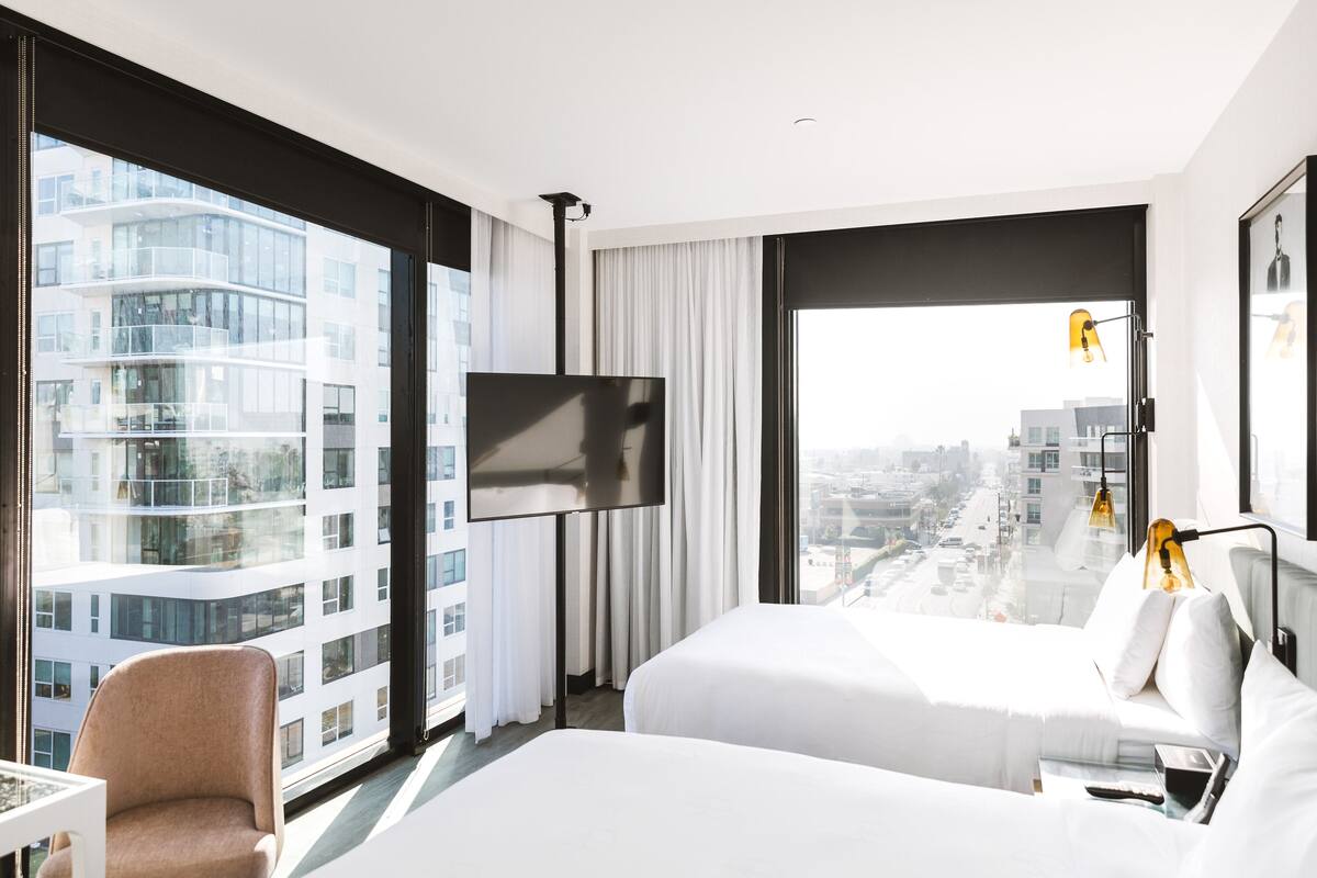 Ein Hotelzimmer mit zwei Betten, einem Fernseher, einem Sessel und einem großen Fenster mit Blick auf die Stadt.