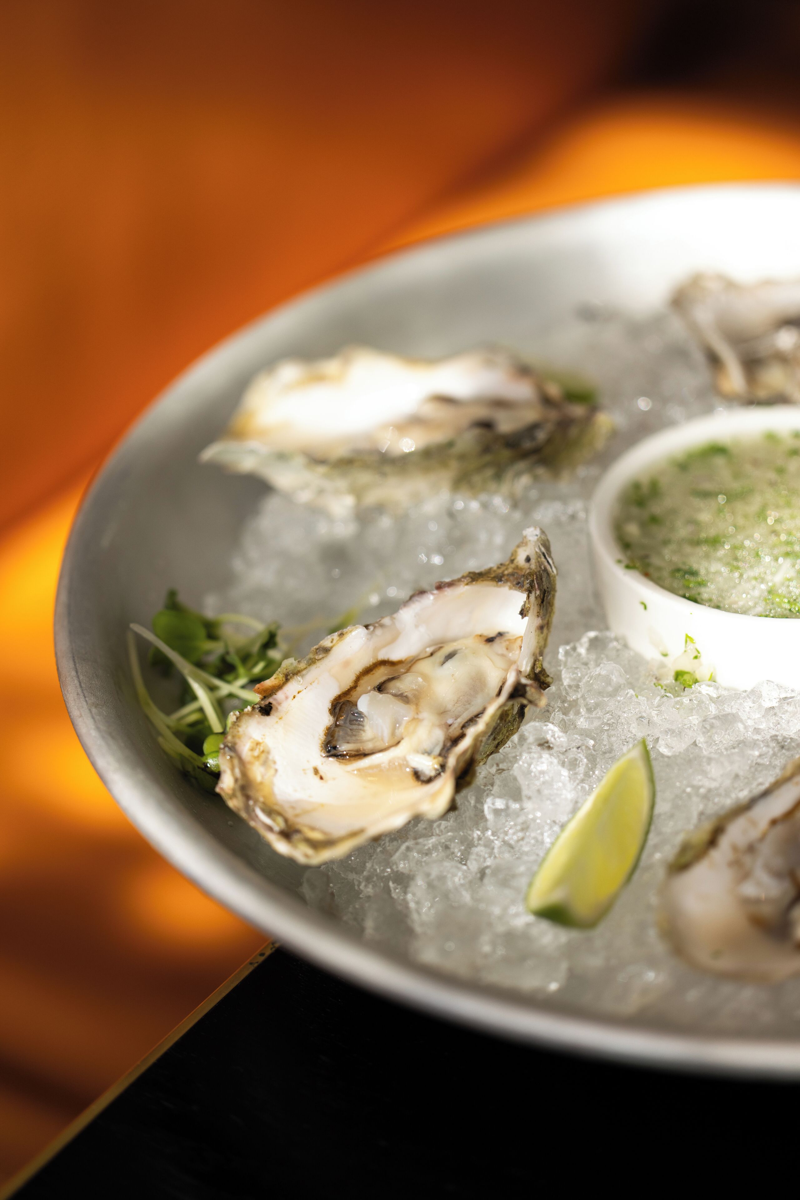 A plate of oysters on ice with a side of green sauce and a lime wedge.