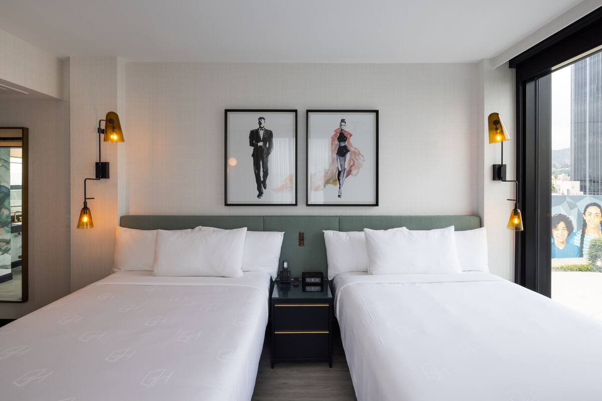 Ein Hotelzimmer mit zwei Betten, einem Nachttisch, einem gerahmten Kunstwerk über den Betten und einem Fenster mit Blick.