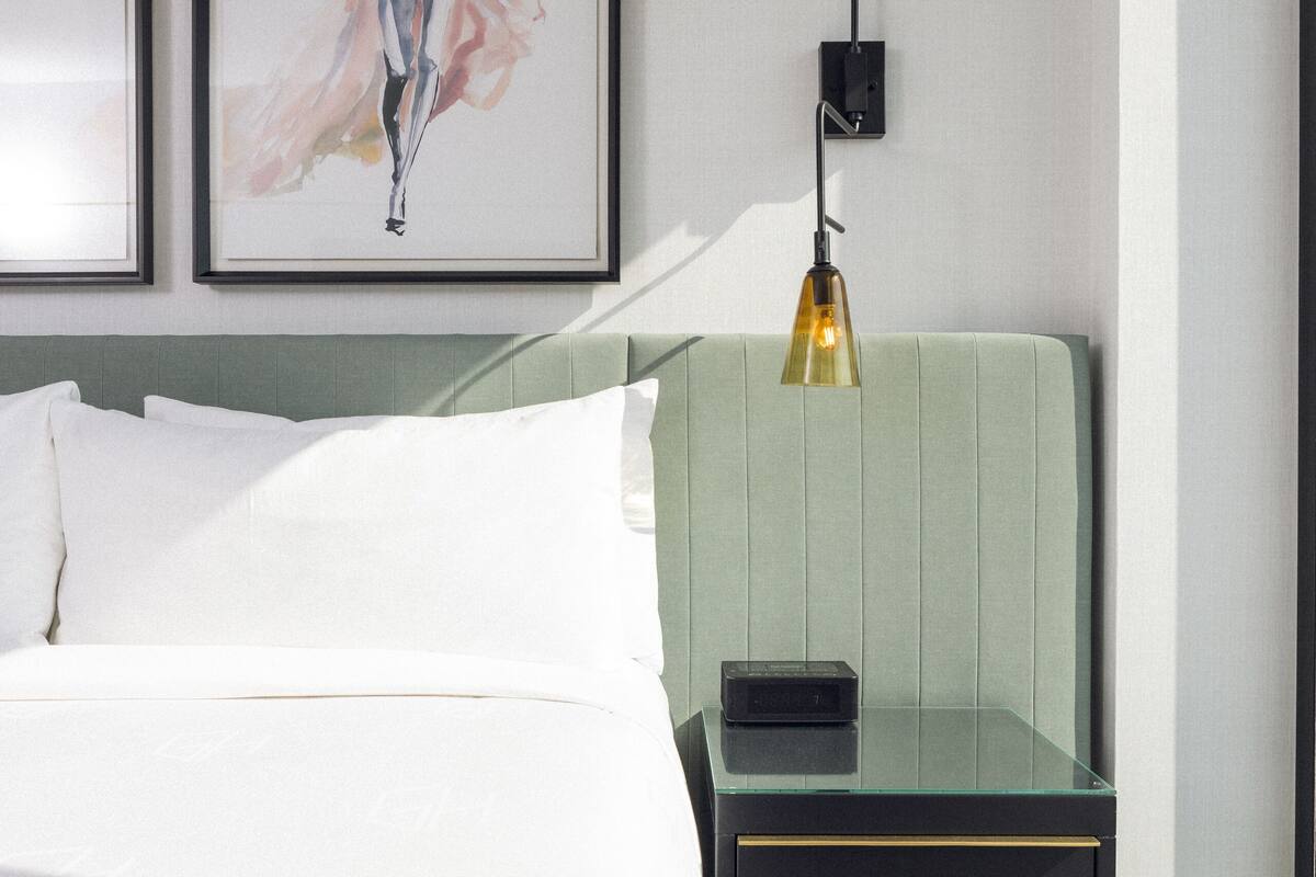 Ein ordentlich bezogenes Bett mit weißen Leinentüchern, einem grünen gepolsterten Kopfteil und einem schwarzen Nachttisch mit Lampe.
