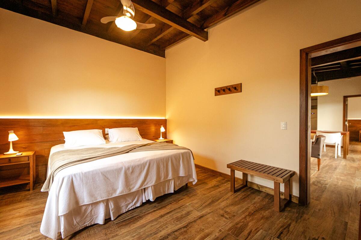 A bedroom with a wooden headboard, a bed with white bedding, a nightstand with a lamp, a wooden bench, and a ceiling fan.
