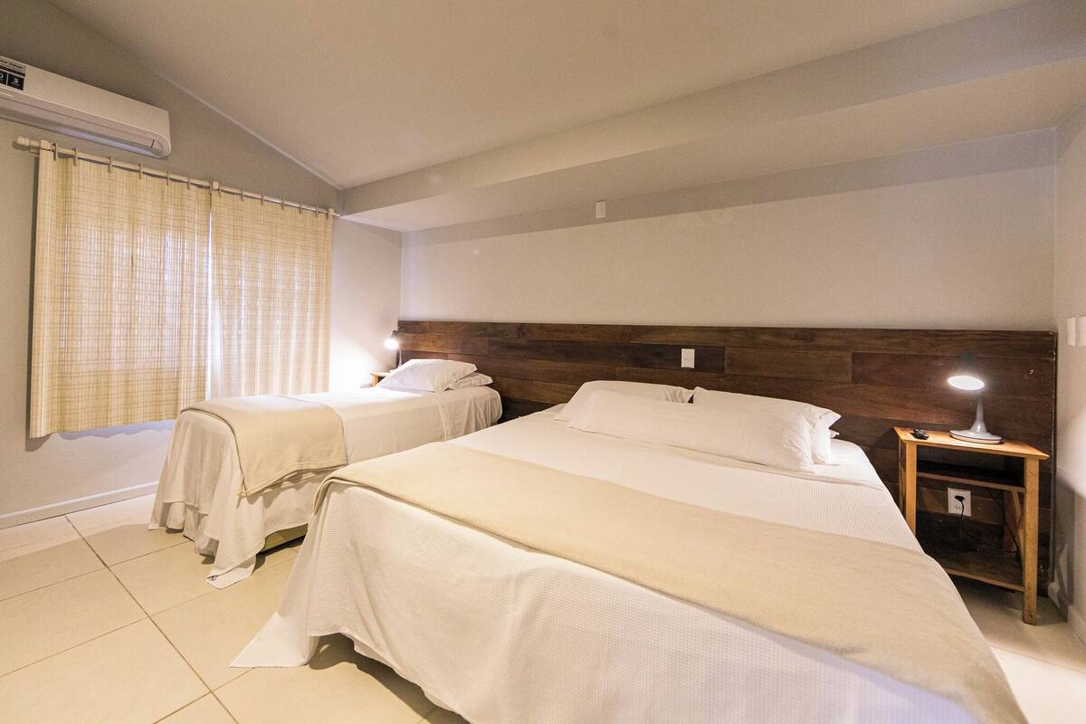 A hotel room with two beds, a wooden headboard, a nightstand with a lamp, and a window with curtains.