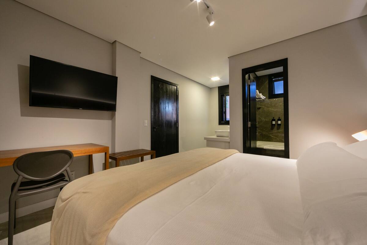 A modern hotel room with a bed, a desk, a chair, a flat-screen TV, a bathroom with a mirror, and a shower.