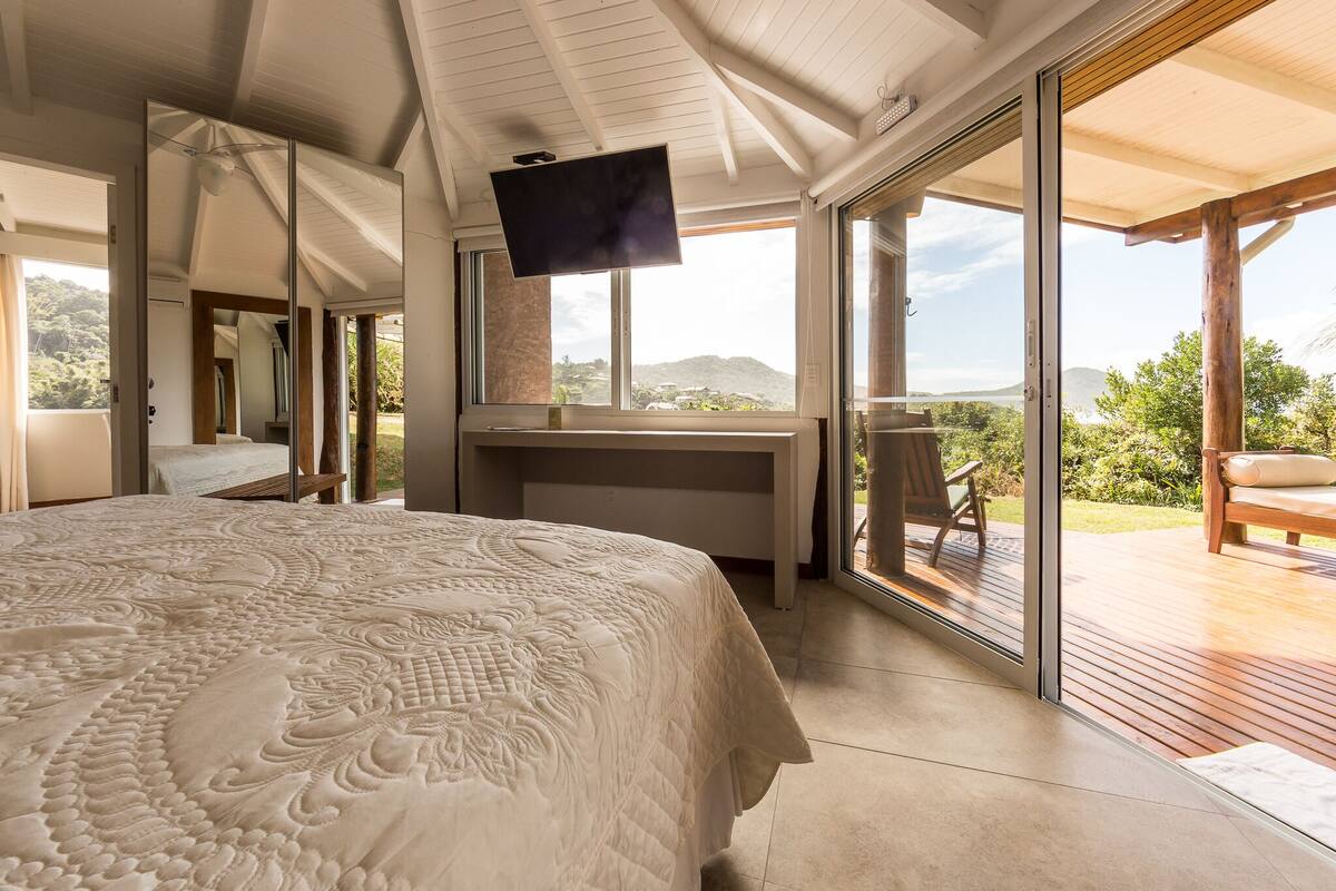 A bedroom with a large bed, a desk, a TV, and a sliding glass door leading to a balcony with a view of the hills.