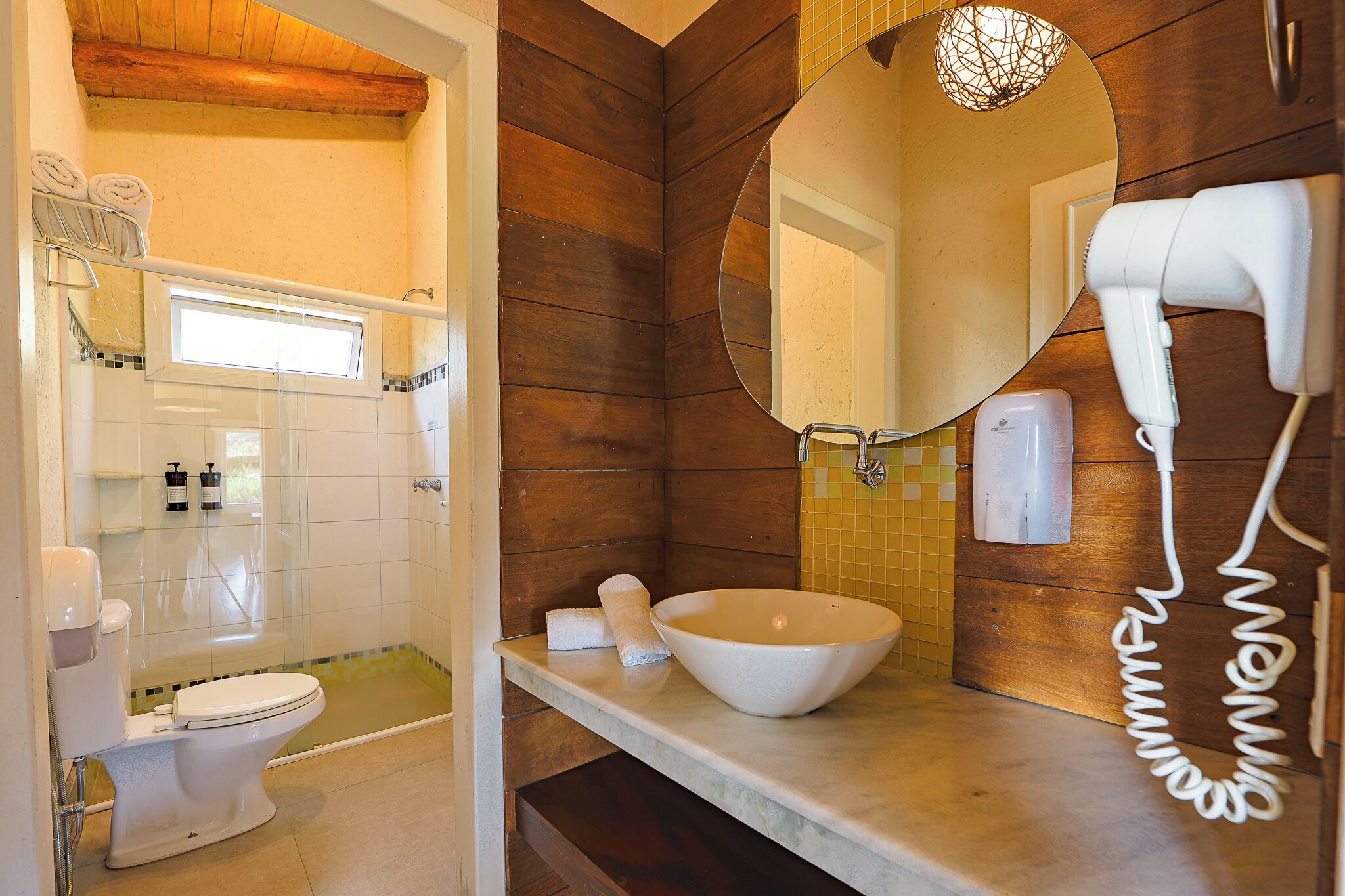 A bathroom with a wooden wall, a large mirror, a white sink, a hairdryer, and a toilet.