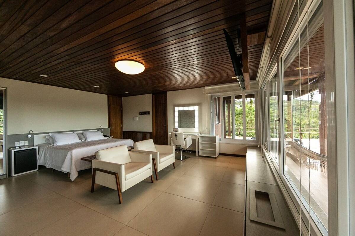 A modern bedroom with a bed, two chairs, a desk, and large windows.