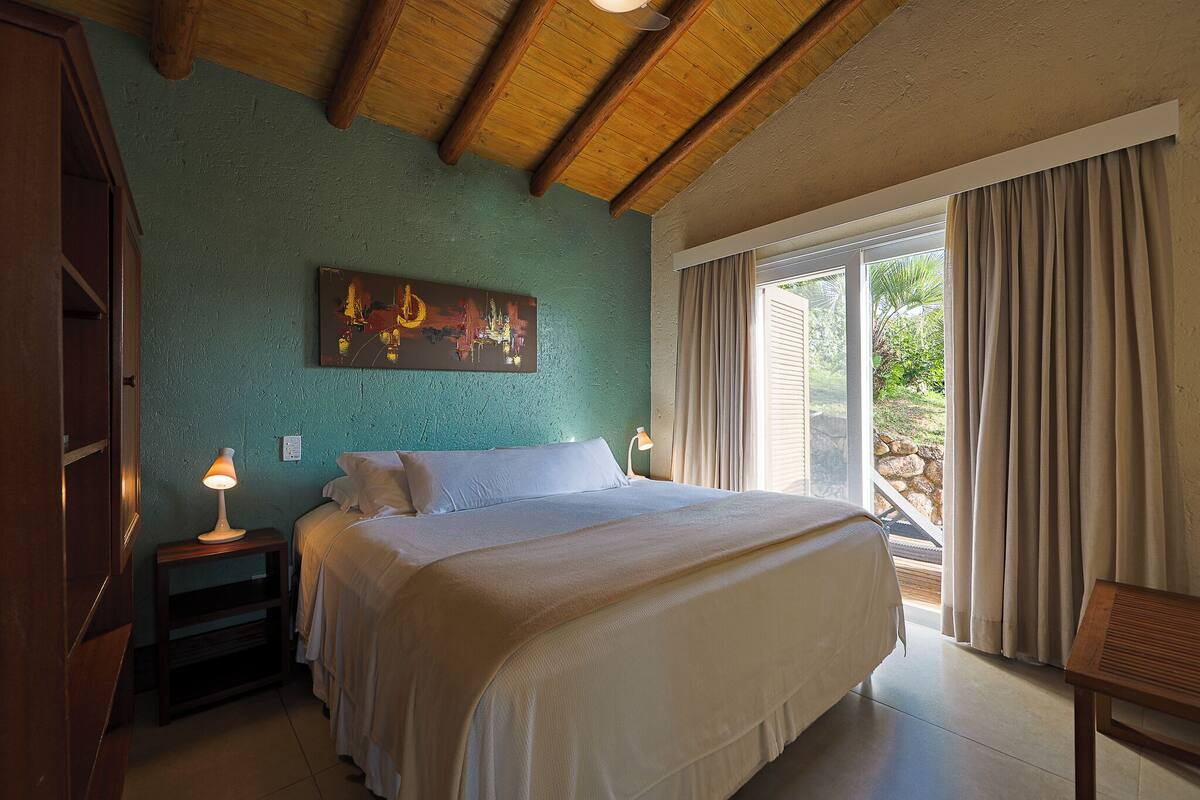 A bedroom with a bed, bedside lamps, a painting, and a view of the outdoors.