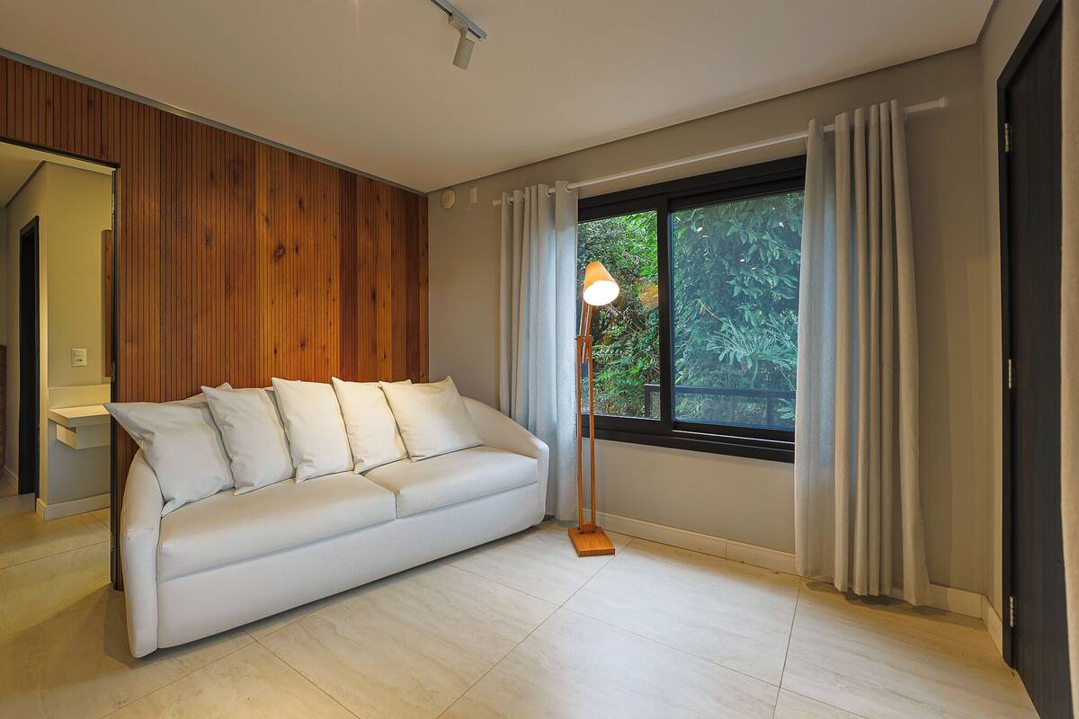 A modern living room with a white sofa, wooden panel walls, a floor lamp, and a large window with curtains.