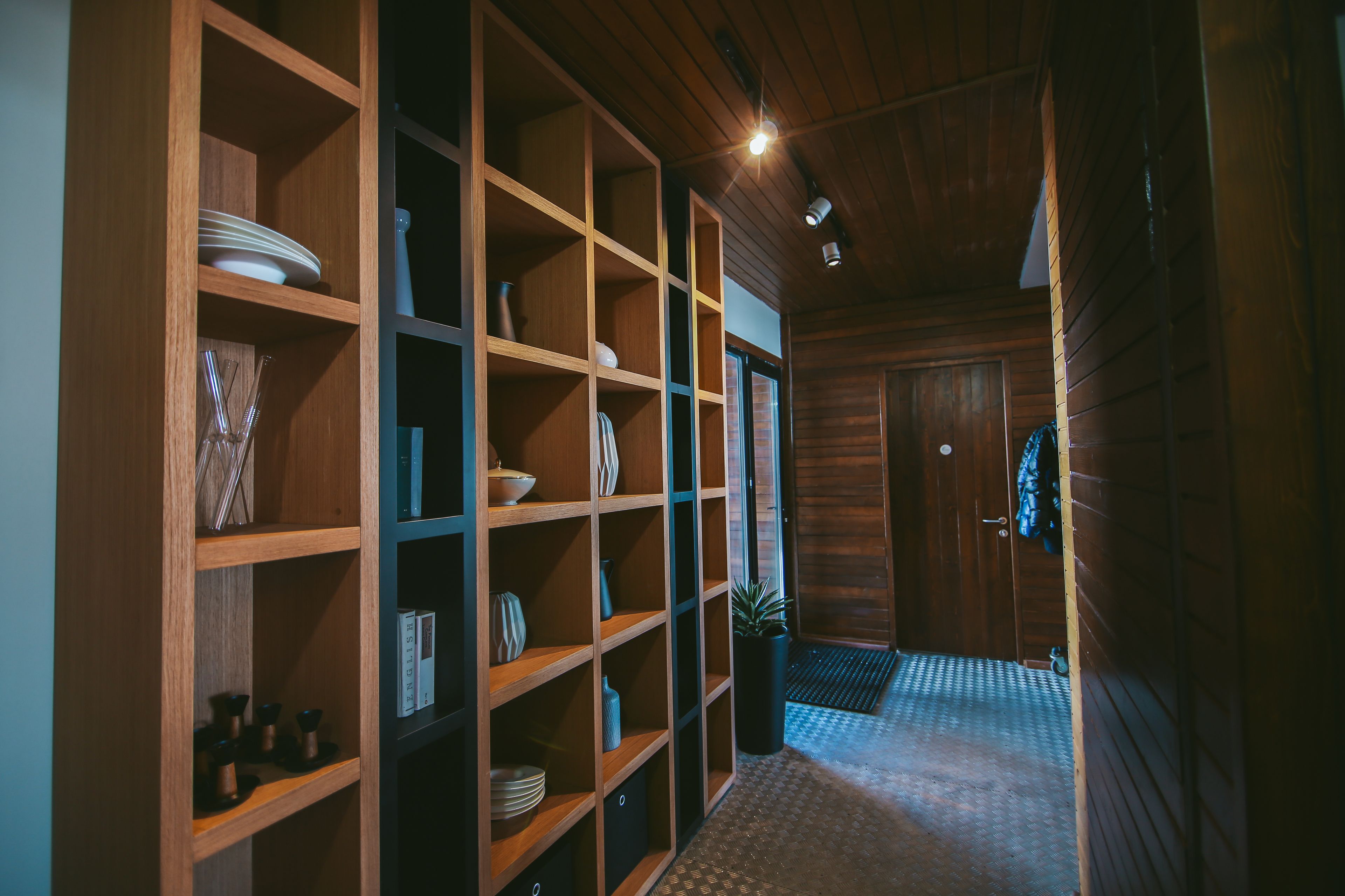 Un interior de madera con un pasillo que conduce a una puerta, estantes con diversos objetos y un suelo con estampado.