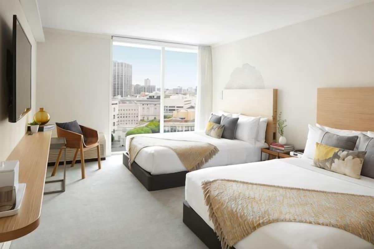 A hotel room with two beds, a desk, a chair, and a large window with a city view.