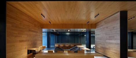 A modern interior with wooden panel walls, a central seating area with tables and chairs, and a visible office space in the background.