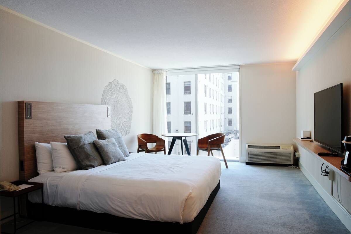 A hotel room with a large bed, a TV, a desk, and a view of buildings through the window.