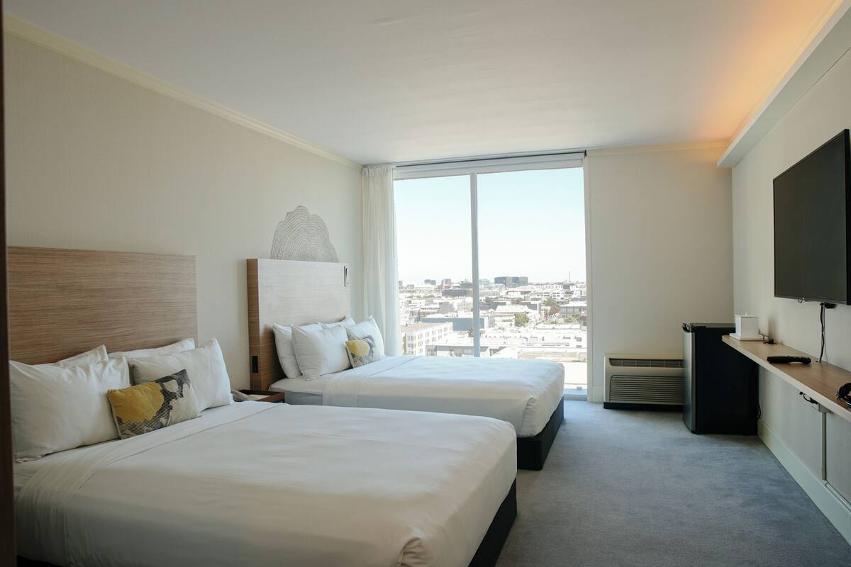 A hotel room with two beds, a large window with a city view, a flat-screen TV, and a desk with a chair.