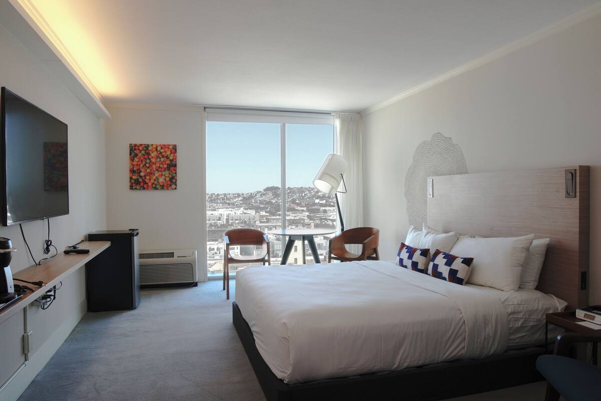 A hotel room with a large bed, a desk with a computer, a chair, a lamp, and a view of the city through a large window.