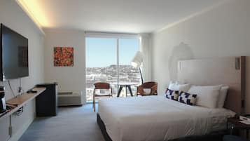 Habitación de hotel con una cama grande, un escritorio con computadora, una silla, una lámpara y vista a la ciudad a través de un ventanal amplio.
