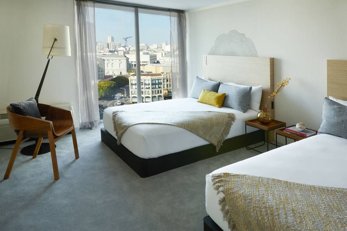 A hotel room with two beds, a desk, a chair, and a view of the cityscape.